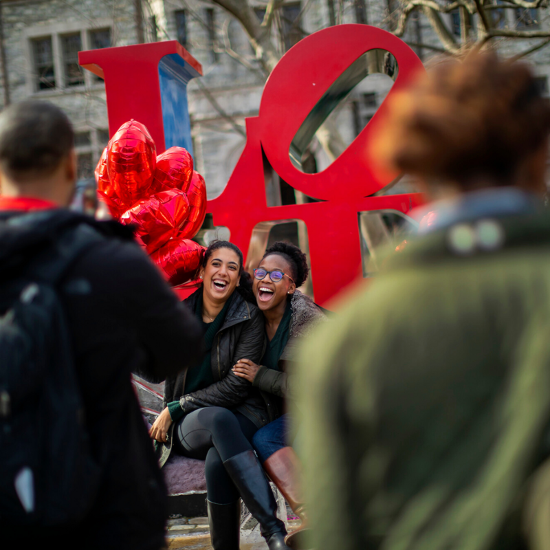 Two people sitting in front of the LOVE sculpture smiling with heart shaped balloons