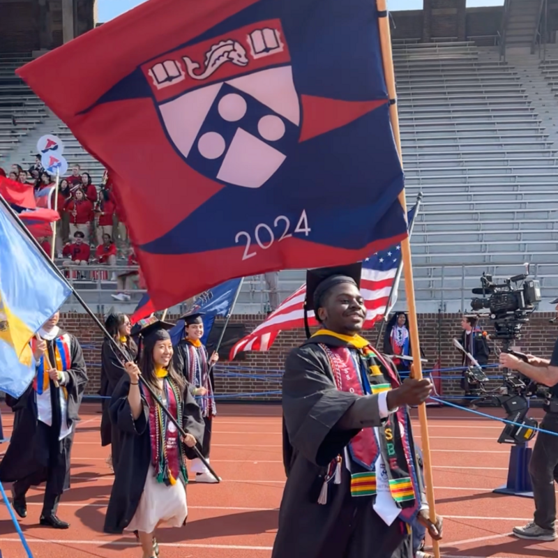 Student wearing academic regalia at commencement carrying a flag featuring the Penn logo and the number 2024